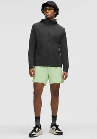 En ung man som bär en svart zip-up jacka, neongula shorts, svart keps, gula solglasögon, svartvita sneakers och svarta mönstrade strumpor medan han står.