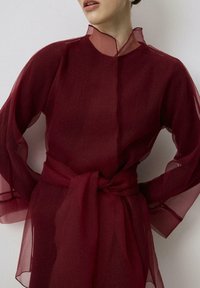 Blusa larga de manga larga transparente color burdeos con cuello alto, cintura elástica y cinturón atado. Tejido texturizado con patrón visible y capas.