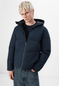 Navy puffer jacket with horizontal seams, front zipper, and a hood. Worn over a dark shirt and paired with faded blue jeans.