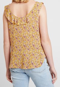 Gul blommig topp med volangkrage, prydd med rosa och vita mönster. Baksidan har en sänkt design, matchad med ljusblå jeans.