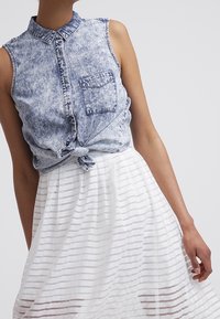 Chemise en denim délavé acide sans manches nouée à la taille, associée à une jupe blanche avec des rayures transparentes horizontales, portée par une personne debout.