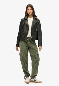 Veste de biker en cuir noir avec des fermetures éclair dorées, t-shirt blanc, pantalon cargo vert olive et baskets blanches avec des accents noirs.