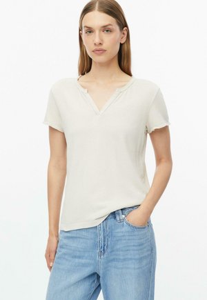 Junge Frau mit glatten hellbraunen Haaren trägt ein off-white kurzärmeliges Oberteil und hellblaue Jeans und steht mit einer Hand in der Tasche.