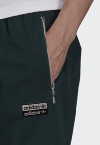 Pantaloni verde scuro con una tasca a zip laterale e una patch con il logo Adidas in bianco e nero, realizzati in tessuto liscio e con un fit standard.