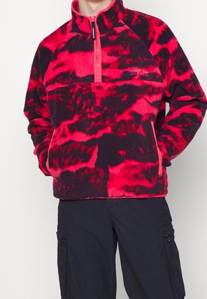 Punainen ja musta tie-dye -kuvioitu fleece-pullover, jossa on puolivetoketju ja kädet taskuissa, yhdistettynä tummiin cargo-housuihin.