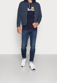 Marinblå bomberjacka över en mörk hoodie med logga, blå denimjeans och vita sportskor. Avslappnad outfit, figurnära stil, få accessoarer.