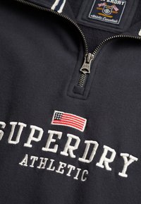 Gros plan sur un sweat-shirt à col zippé court bleu marine foncé avec "SUPERDRY ATHLETIC" brodé en blanc et un petit drapeau américain au-dessus du texte.