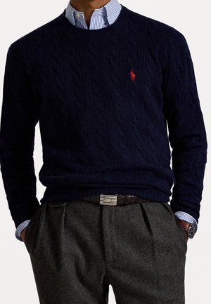 Man wearing navy cable-knit sweater with red logo, blue striped collared shirt, gray pleated trousers, and black belt with silver buckle.