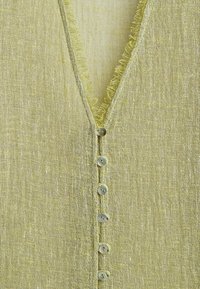 Tissu texturé vert clair avec un col en V et sept petits boutons ronds au centre, présentant des bords effilochés le long de l'encolure.