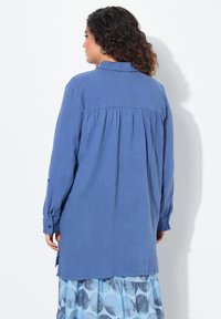 Blusa blu a maniche lunghe con finitura testurizzata, caratterizzata da una piega sul retro e polsini con bottoni. Abbinata a una gonna blu a fantasia.