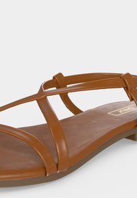 Καφέ επίπεδη σανδάλι με λεπτά, λείο δερμάτινα λουριά που διασχίζουν το πόδι και στρογγυλεμένη σόλα.