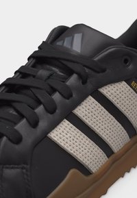 Must ja beež värvi Adidas toss, millel on mustad paelad, küljel kolm beeži võrgusilma triipu ja kummist värvi tallaosa.