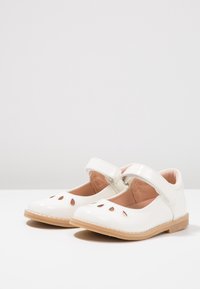 Białe lakierowane buty z zaokrąglonym noskiem, regulowanym paskiem i wycięciami z przodu. Charakteryzują się brązową gumową podeszwą.