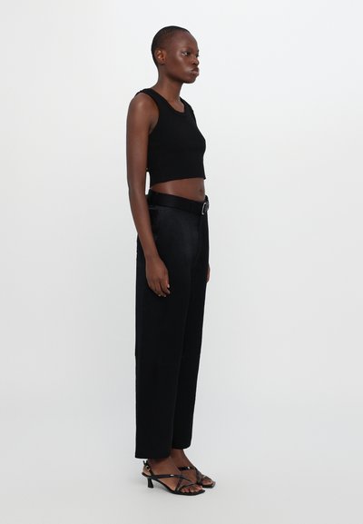 Zwarte cropped tanktop, hoge zwarte broek met riem en zwarte sandalen met straps. Soepele texturen en slank ontwerp. Zijkant profielweergave.