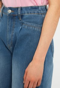 Κοντινό πλάνο ενός ατόμου που φορά μπλε τζιν και ροζ μπλούζα, με το δεξί χέρι χαλαρό στο πλάι.