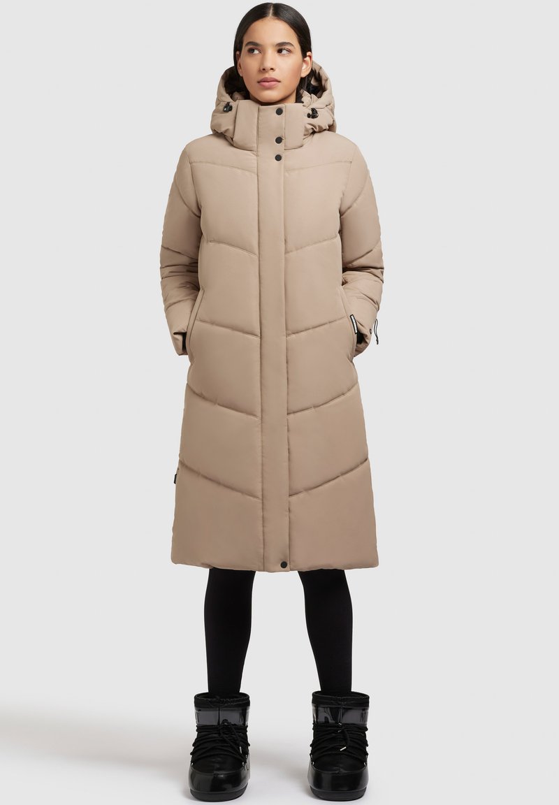 Beige gepolsterter langer Mantel mit Kapuze, vertikaler Steppung, vorderen Knöpfen und Reißverschlusstaschen an der Seite. Kombiniert mit schwarzen Stiefeln.