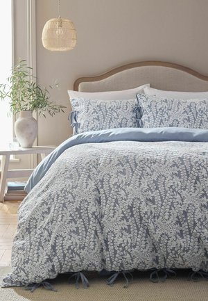 Bed with blue and white floral patterned bedding, beige upholstered headboard, hanging wicker lamp, and a side table with a vase of green branches.