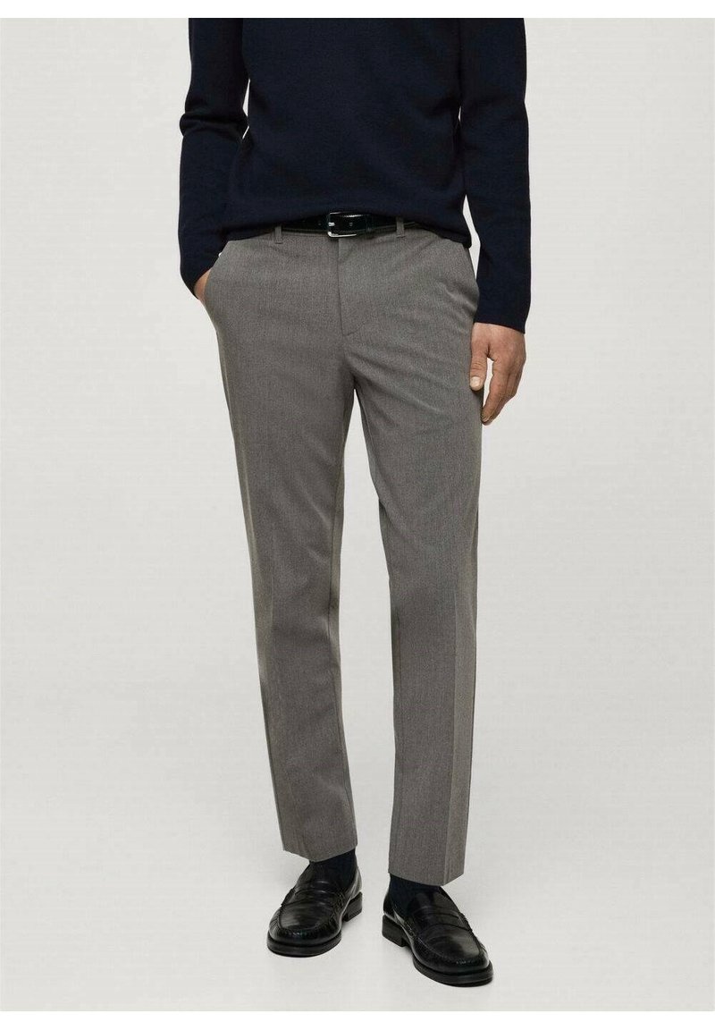 Homme portant un pantalon gris sur mesure, une ceinture noire, des mocassins noirs et un haut sombre à manches longues, debout les mains partiellement dans les poches.