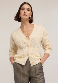 Krémový pletený cardigan s volným vzorem, s výstřihem do V, předními kapsami a zapínáním na knoflík. Hladké manžety a spodní lem.