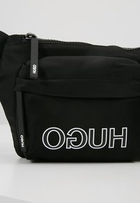 Borsa a cintura in tessuto nero con chiusura a zip, dotata di tasca frontale e audace logo bianco "HUGO". Texture liscia, design minimalista.