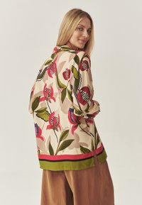 Vrouw met blond haar, gekleed in een beige blouse met lange mouwen met grote rode en groene bloemenpatronen, gecombineerd met bruine losse broeken, kijkt om.