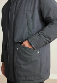 Mann trägt einen dunkelgrauen Mantel mit einer Hand in der großen Fronttasche, die Druckknopfdetails und eine vertikale Brusttasche zeigt.