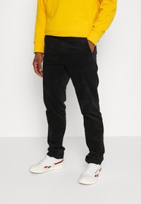 Svarta kordflamsbyxor med en strukturerad yta, bärs med en gul sweatshirt och vita sneakers med röda och blå detaljer.