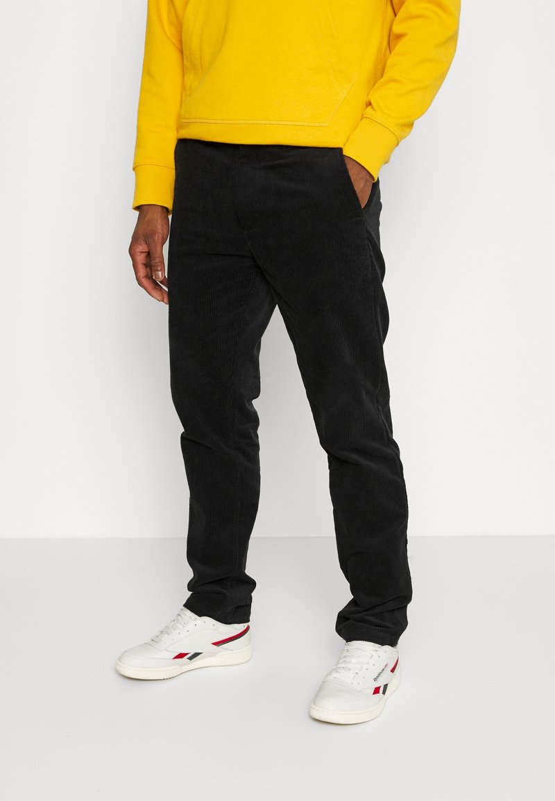 Svarta kordflamsbyxor med en strukturerad yta, bärs med en gul sweatshirt och vita sneakers med röda och blå detaljer.