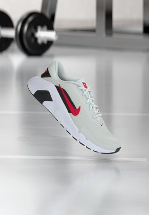 Hellgrauer Nike Laufschuh mit rotem Swoosh und weißer Sohle, schwebt über reflektierendem Fitnessboden, Gewichthebergeräte im Hintergrund verschwommen.