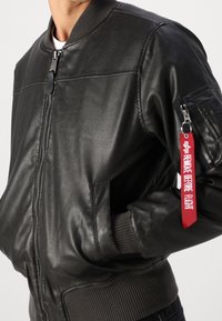 Černá kožená bomber bunda s žebrovaným límcem a manžetami, bočními kapsami na zip a červeným štítkem "ODSTRANIT PŘED ODBOČENÍM" na rukávu.