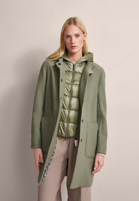 Grüner Wollmischungs-Mantel mit taillierter Passform, zwei Fronttaschen und Knopfverschluss über einer hellgrünen Daunenjacke mit Kapuze.