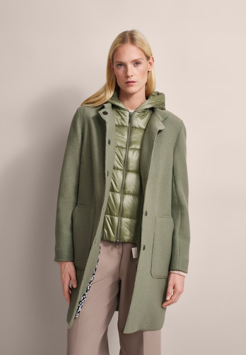 Grüner Wollmischungs-Mantel mit taillierter Passform, zwei Fronttaschen und Knopfverschluss über einer hellgrünen Daunenjacke mit Kapuze.