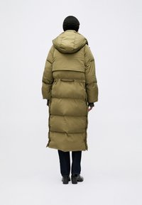 Duga maslinasto zeleni puffer kaput s kapuljačom, s naglašenim strukom, prednjim džepovima i kviltanim uzorkom. Nosi se preko tamnih traperica i čizama.