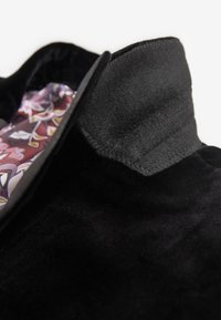 Giacca in suede nera con colletto alto. L'interno presenta una fodera colorata e a fantasia. Design in tessuto strutturato. Dettagli di cucitura su misura.