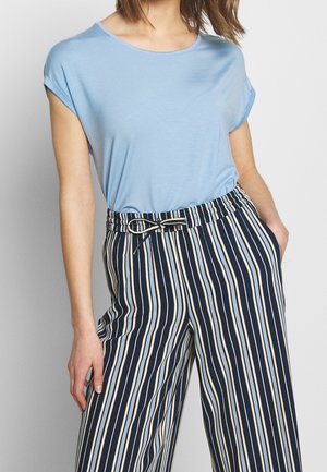 Femme portant un haut bleu clair à manches courtes rentrée dans un pantalon taille haute à rayures verticales bleu marine, blanc et beige avec une ceinture à la taille.