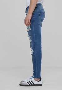 Középkék farmer skinny farmernadrág, a combok mentén kopott szakítással. Fehér sportcipővel párosítva, fekete és szürke díszítéssel.