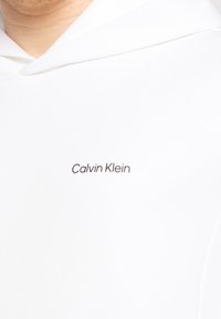 Witte katoenen hoodie met een eenvoudig, modern ontwerp en een klein bruin 'Calvin Klein' logo aan de linkerkant van de borst.