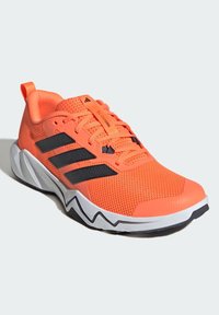 Jasně oranžová atletická obuv s prodyšným svrškem z materiálu mesh, černým designem se třemi pruhy a bílou podešví se zigzag vzorem. Má polstrovaný límec a šněrovací systém.