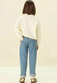 Sweatshirt blanc surdimensionné, jeans bleus avec broderie en forme de cœur jaune, poignets retroussés, et chaussures marron avec des chaussettes ; vu de derrière.