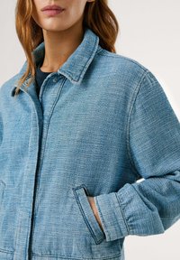 Blue denim jacket with a textured finish, featuring a shirt collar, button closure, and side pockets. Soft fabric and relaxed fit.