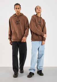 Due persone indossano felpe marroni con grafiche di falene nere, una in jeans neri e l'altra in jeans azzurri chiari, entrambe con scarpe nere stringate.