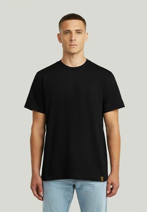 Man met kort haar, gekleed in een effen zwart T-shirt met korte mouwen en lichtblauwe spijkerbroek, staand tegen een eenvoudige lichte achtergrond.