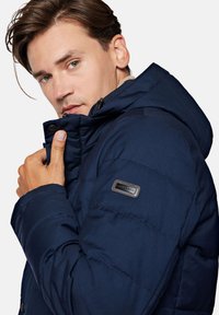 Granatowa pikowana kurtka z kapturem, z zamkiem błyskawicznym, akcentami guzikowymi i naszywką marki na lewym ramieniu. Miękka tekstura.