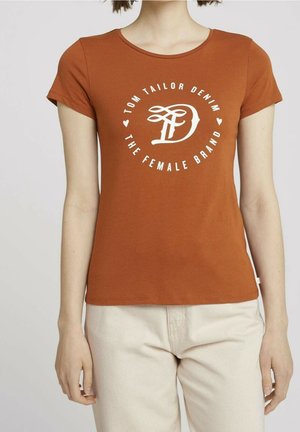 Femme portant un T-shirt à manches courtes couleur rouille avec le texte "Tom Tailor Denim The Female Brand" et un pantalon beige.
