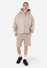 Beige zip-up hoodie med en frontlomme, parret med matchende beige shorts. Båret med sorte sneakers og hvide sokker, som har logoaccenter.