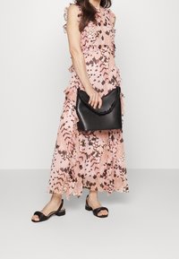 Musta nahkalaukku, jossa on rypistetty kahva, käytetty vaaleanpunaisen ja beigeen kuvioidun mekon kanssa, jonka helmoissa on frilloja, sekä mustien nilkkaremmillä varustettujen sandaalien kanssa.