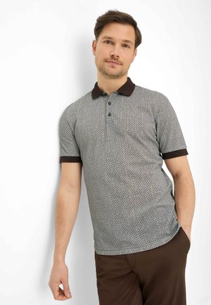 Mand, der læner sig op ad en hvid væg iført en kortærmet polo med et lille ternet mønster og brun krave, kombineret med brune bukser.