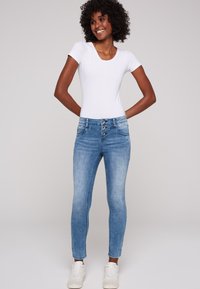 Jeans skinny de mezclilla azul con cintura alta, que cuentan con tres botones, cinco bolsillos y un lavado ligeramente deslavado. Combinados con una camiseta blanca.
