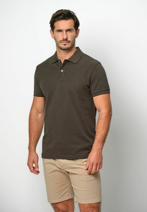 Bruin poloshirt met kraag en korte mouwen, voorzien van een knoopsluiting met drie knopen. Draagt beige korte broek, tegen een neutrale achtergrond.