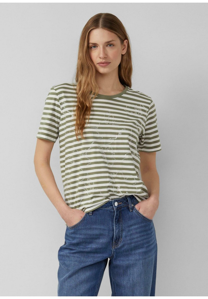 Vrouw met lang lichtbruin haar, gekleed in een groen-wit gestreept shirt met korte mouwen en een blauwe spijkerbroek, met handen in de zakken tegen een effen grijze achtergrond.